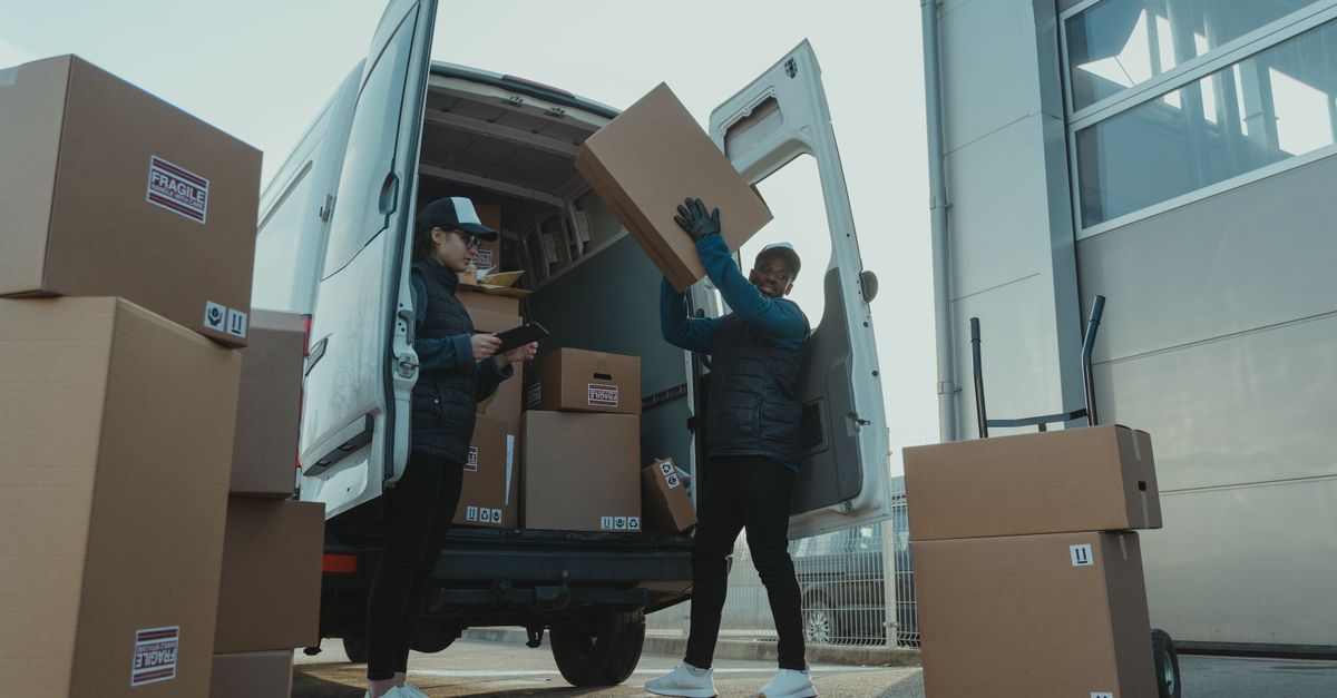 Workers loading boxes into a delivery van at a warehouse facility outdoors.